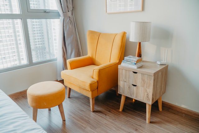 A yellow armchair and a stool next to a bedside table by a window