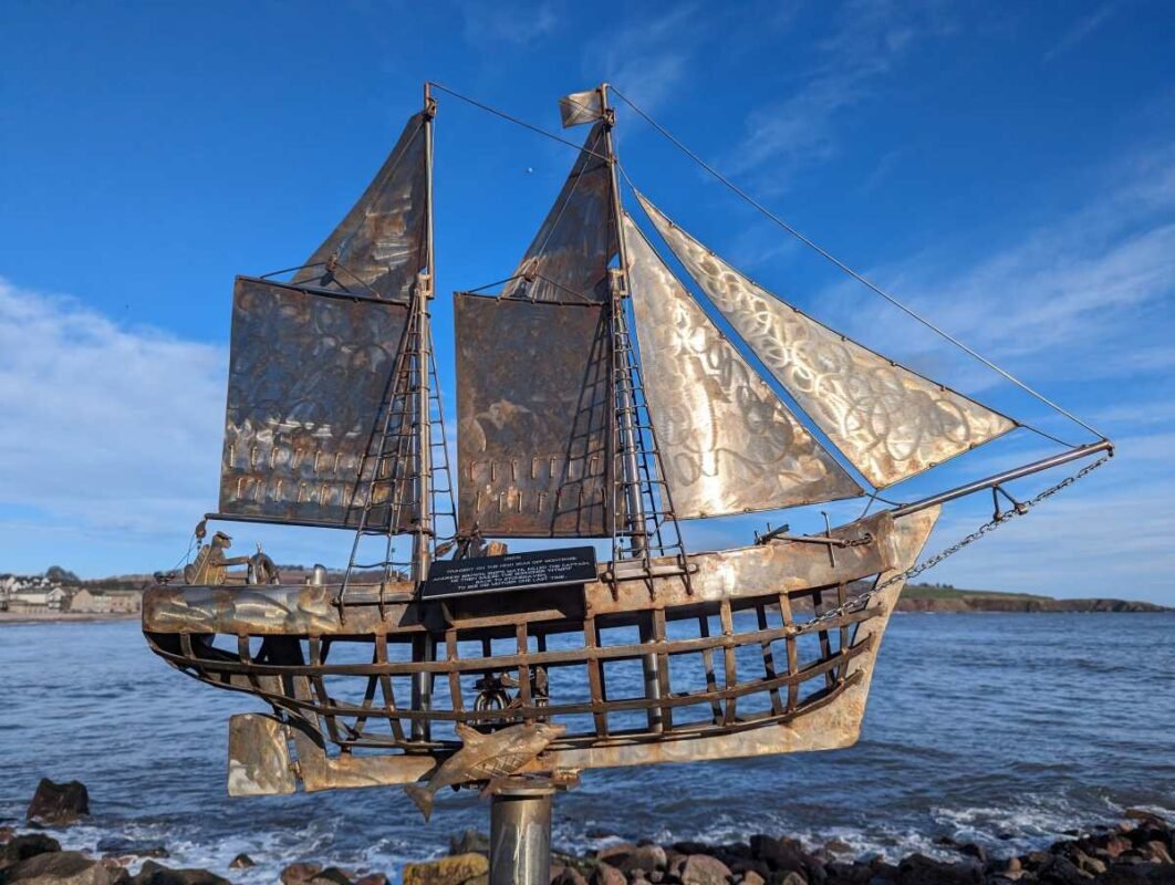 Metal sailboat sculpture by the sea under blue skies.