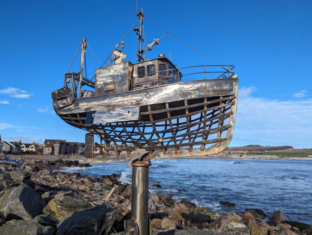 Metal boat sculpture near rocky shoreline under clear blue sky.
