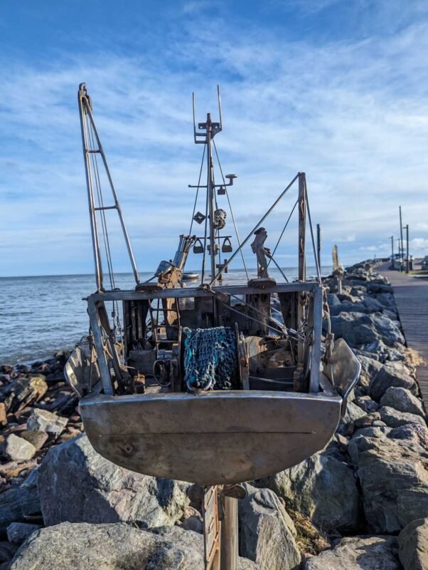 Metal sculpture of a boat on rocky shoreline.