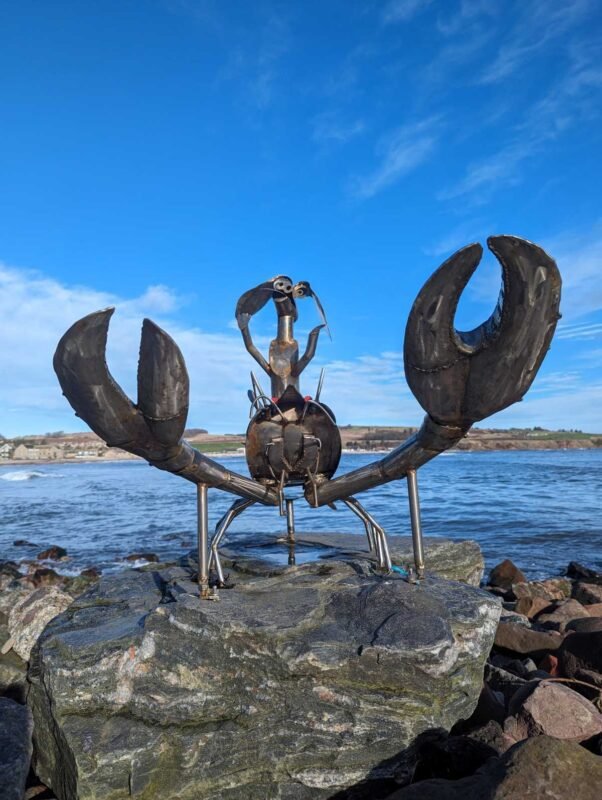 Metal crab sculpture by the sea on a rocky shore.