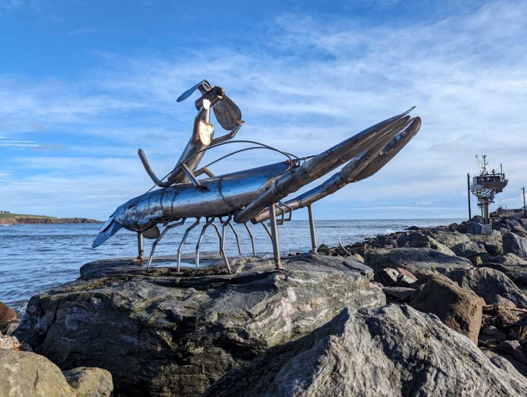 Steel lobster sculpture by the ocean on rocky shore.
