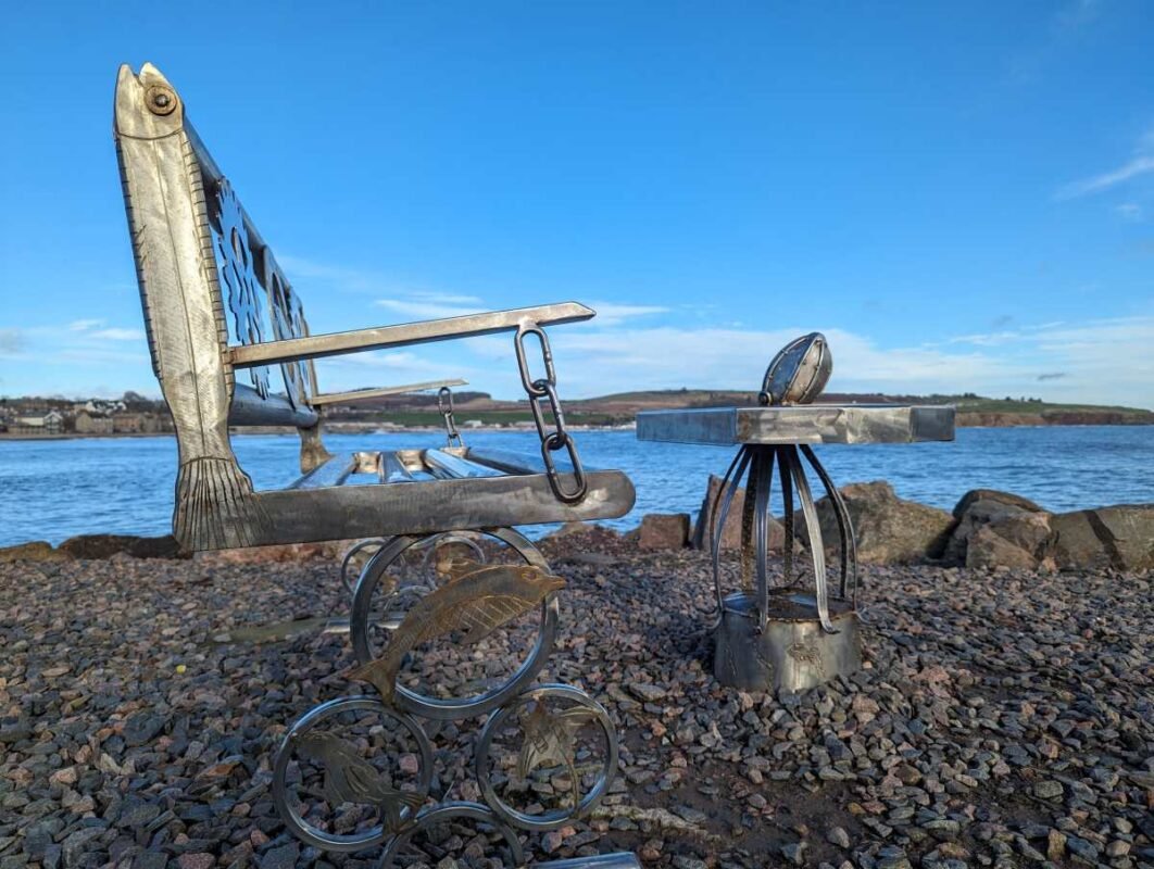 Metal fish-themed bench by a scenic waterfront.