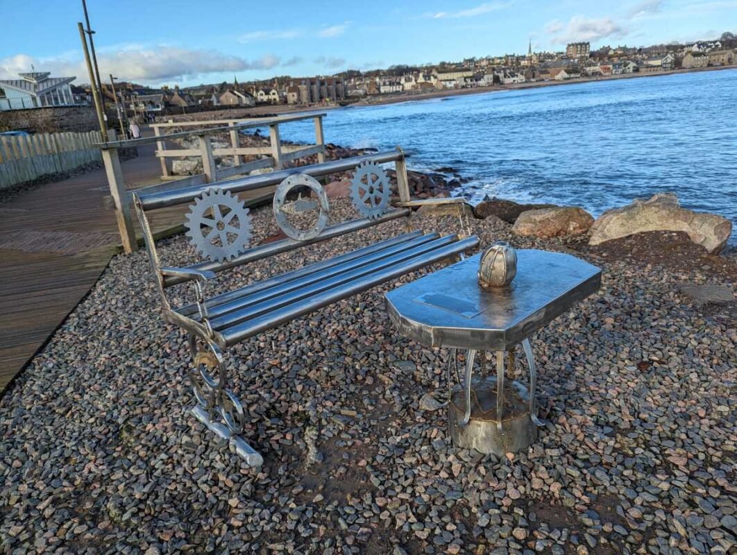 Seaside metal bench with gear design near waterfront.