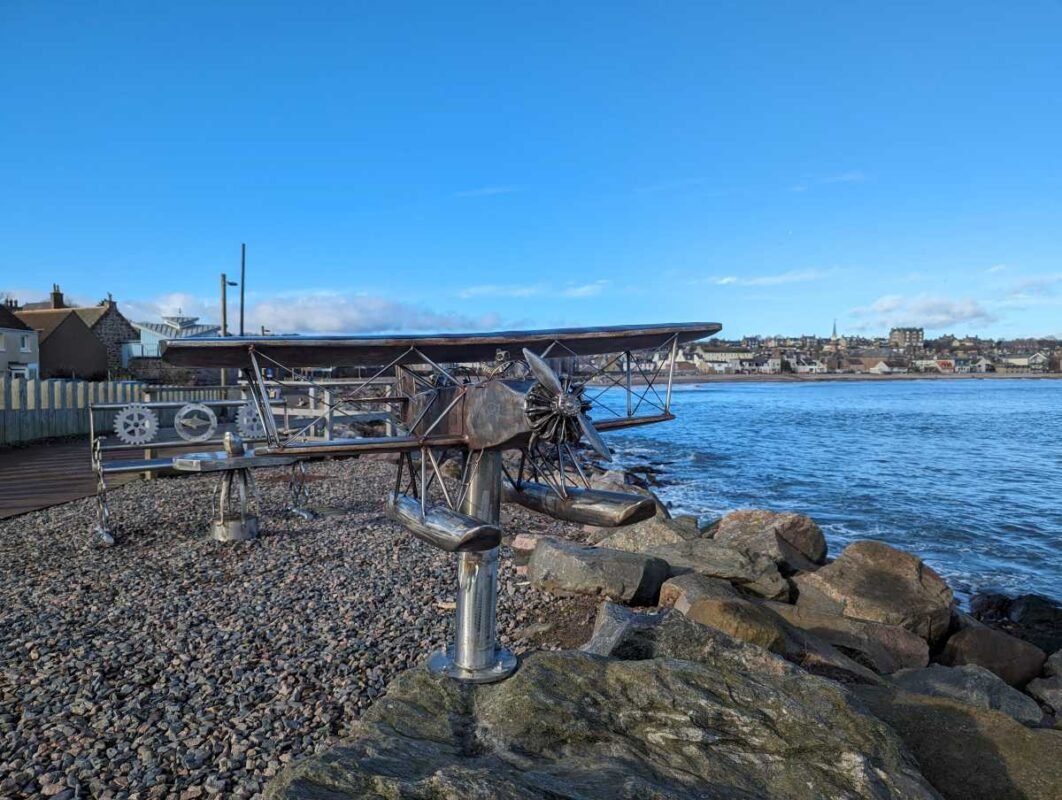 Metal plane sculpture by the sea with coastal town backdrop.