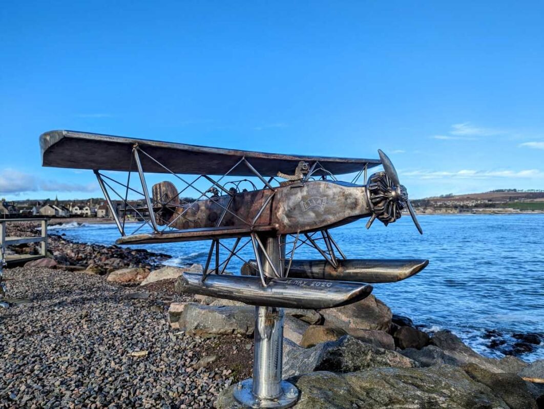 Metal airplane sculpture by the sea on a rocky shore.